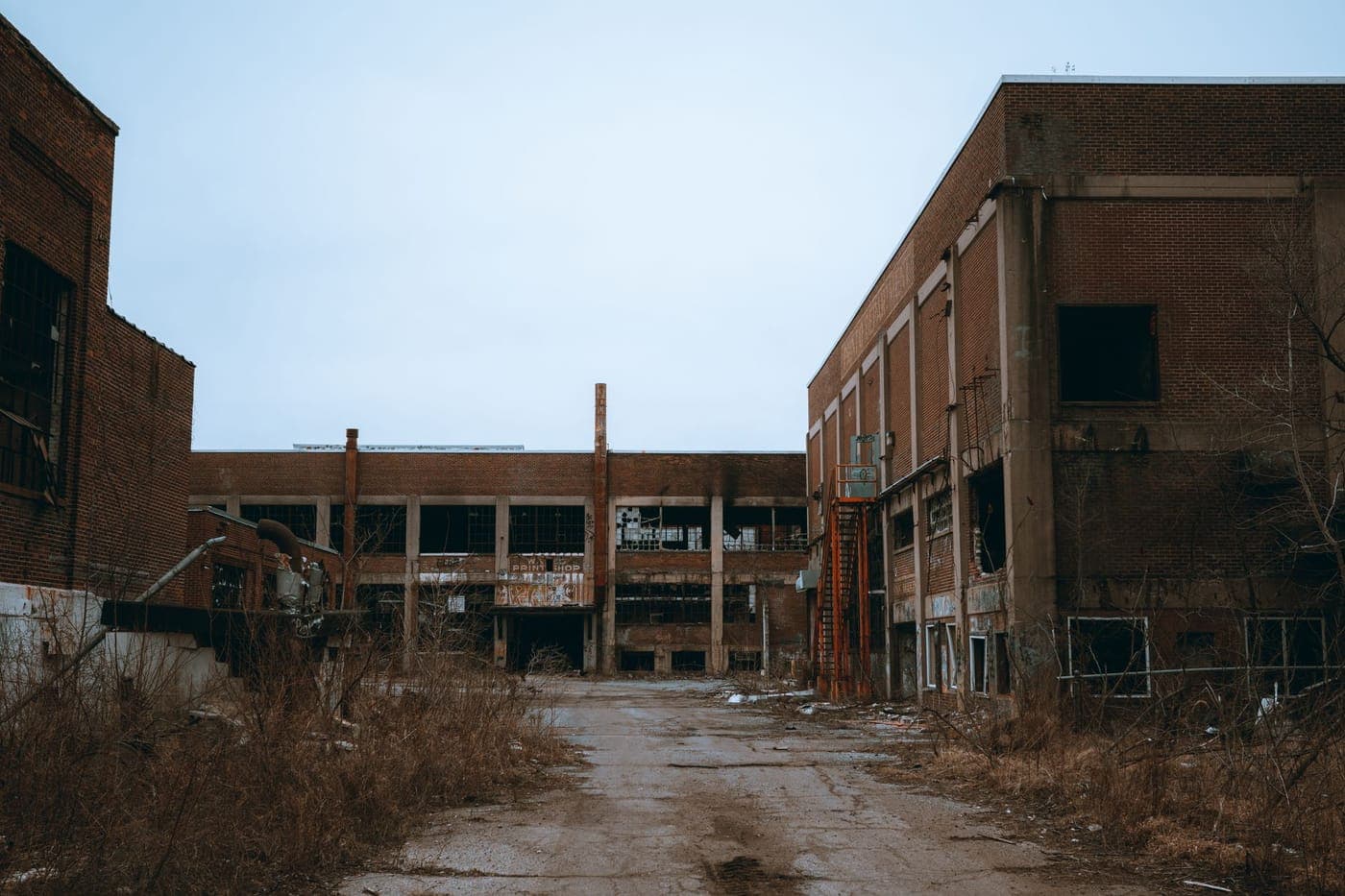 Abandoned brick factory buildings and an overgrown yard under a grey sky