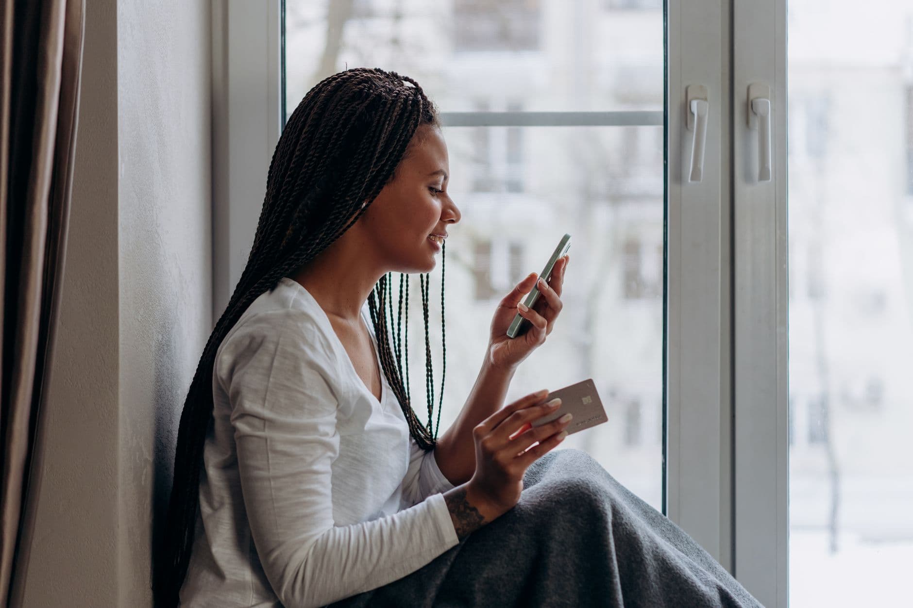 Woman sitting by a window shopping online with her phone and credit card.